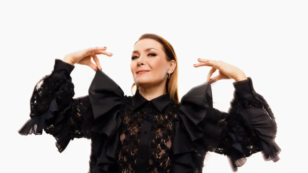 Irina mustassa pitsikoristeisessa asussa, kädet kohotettuina, vaaleaa taustaa vasten.