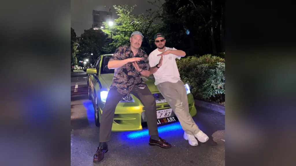 Kaksi miestä poseeraa vihreän auton edessä yöaikaan. Autossa on sininen alavalaistus.